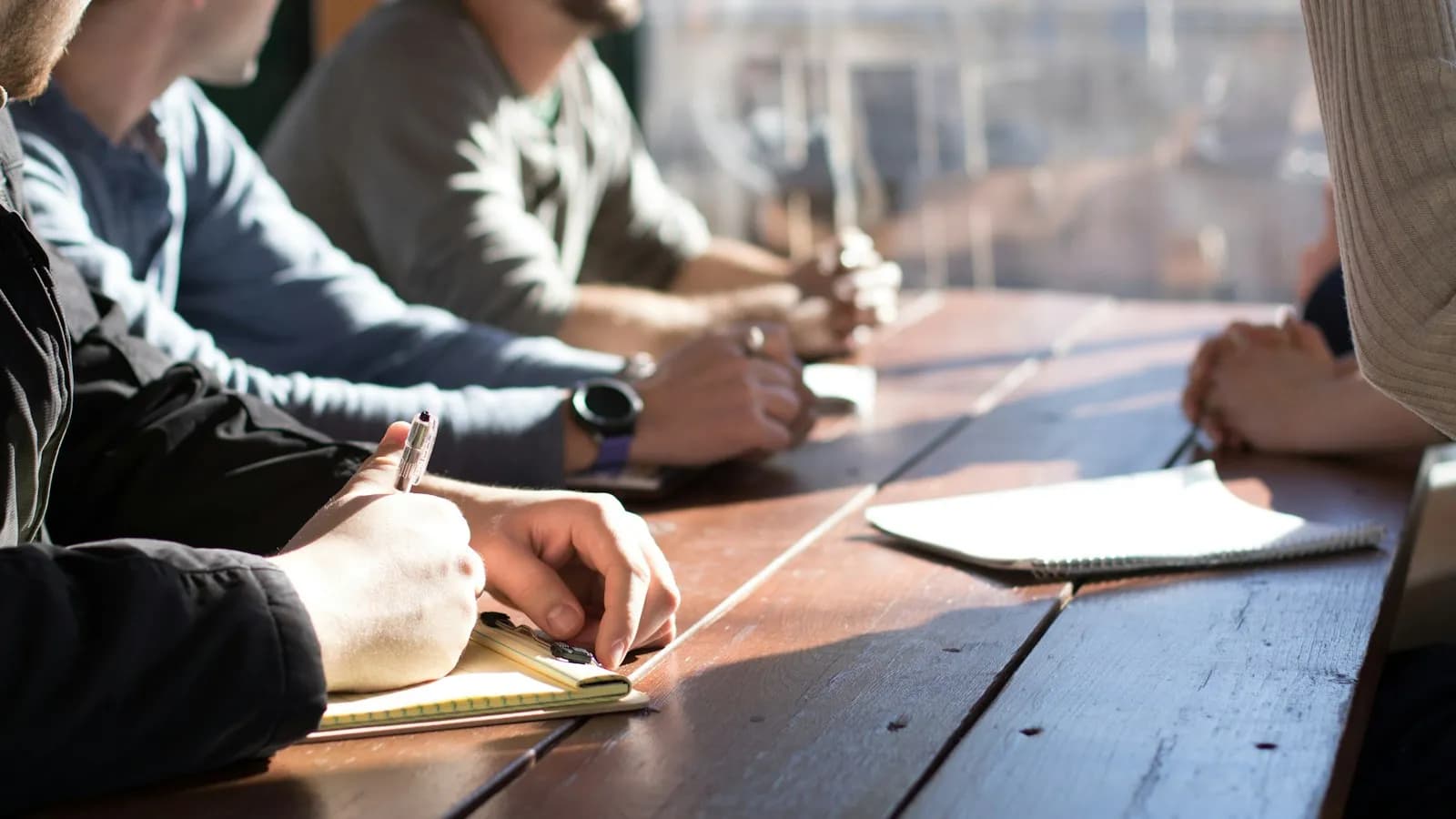 People sitting around a wooden table in a meeting, one person taking notes
