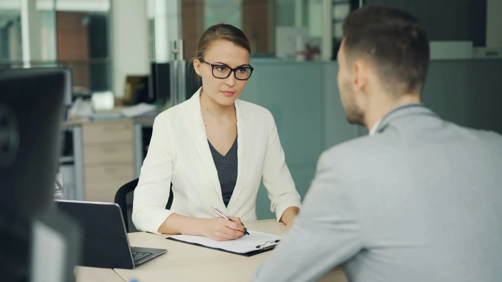 An interviewer in glasses talking with a candidate across an office desk