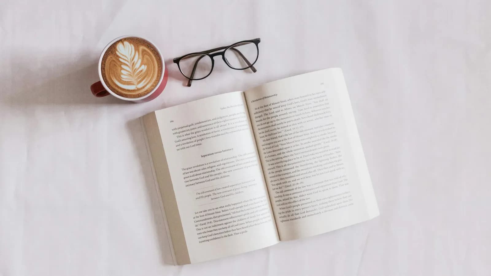 An open book on a white table next to a cup of coffee and a pair of glasses