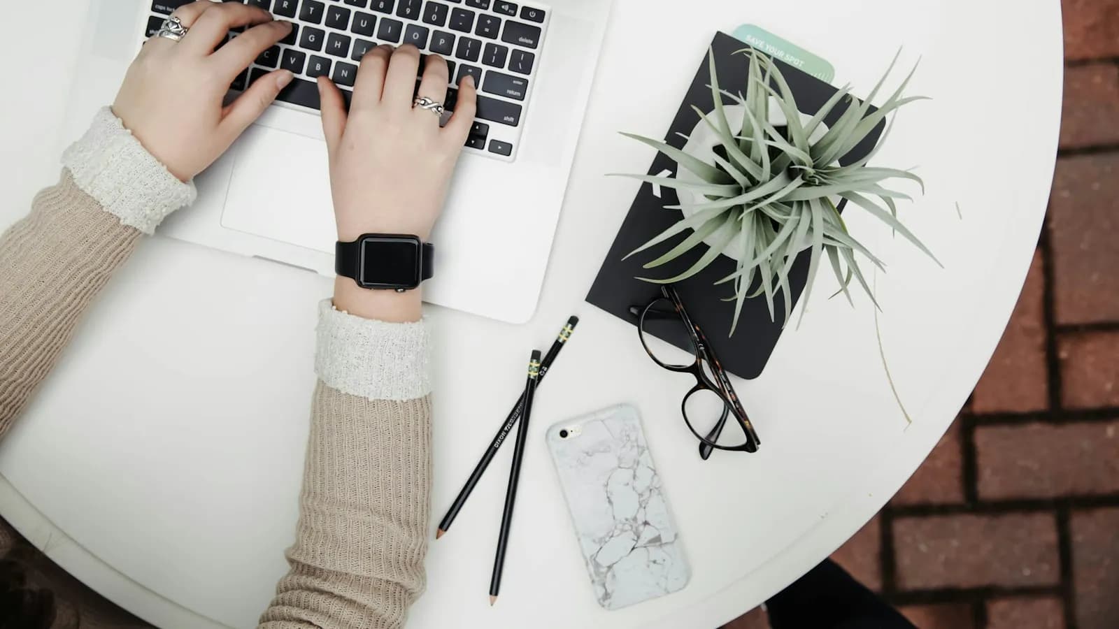 A person typing on a laptop at a white table with a plant, glasses and a phone beside it