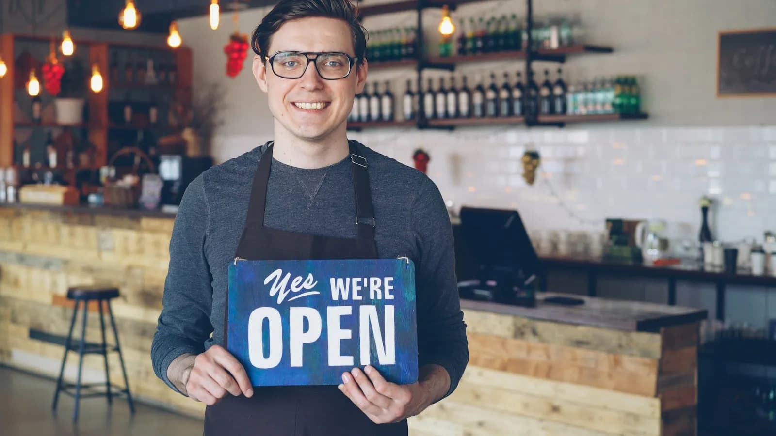 A smiling cafe owner in an apron holding a sign that says Yes we're open