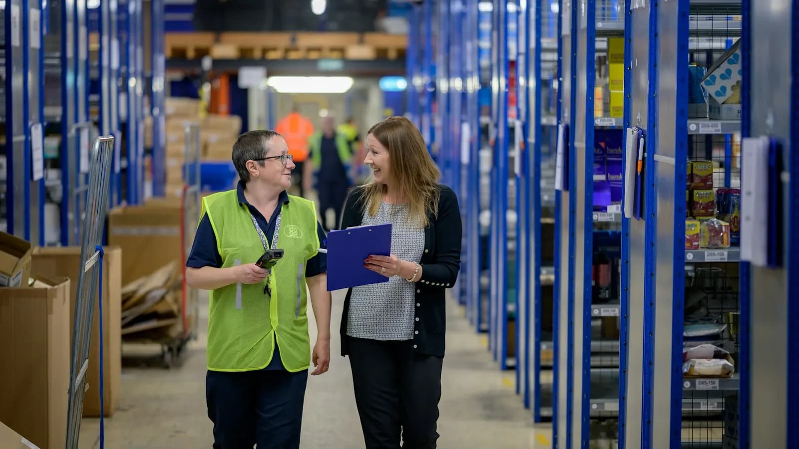 Two staff members walking down a store aisle lined with shelves of stock, one holding a clipboard