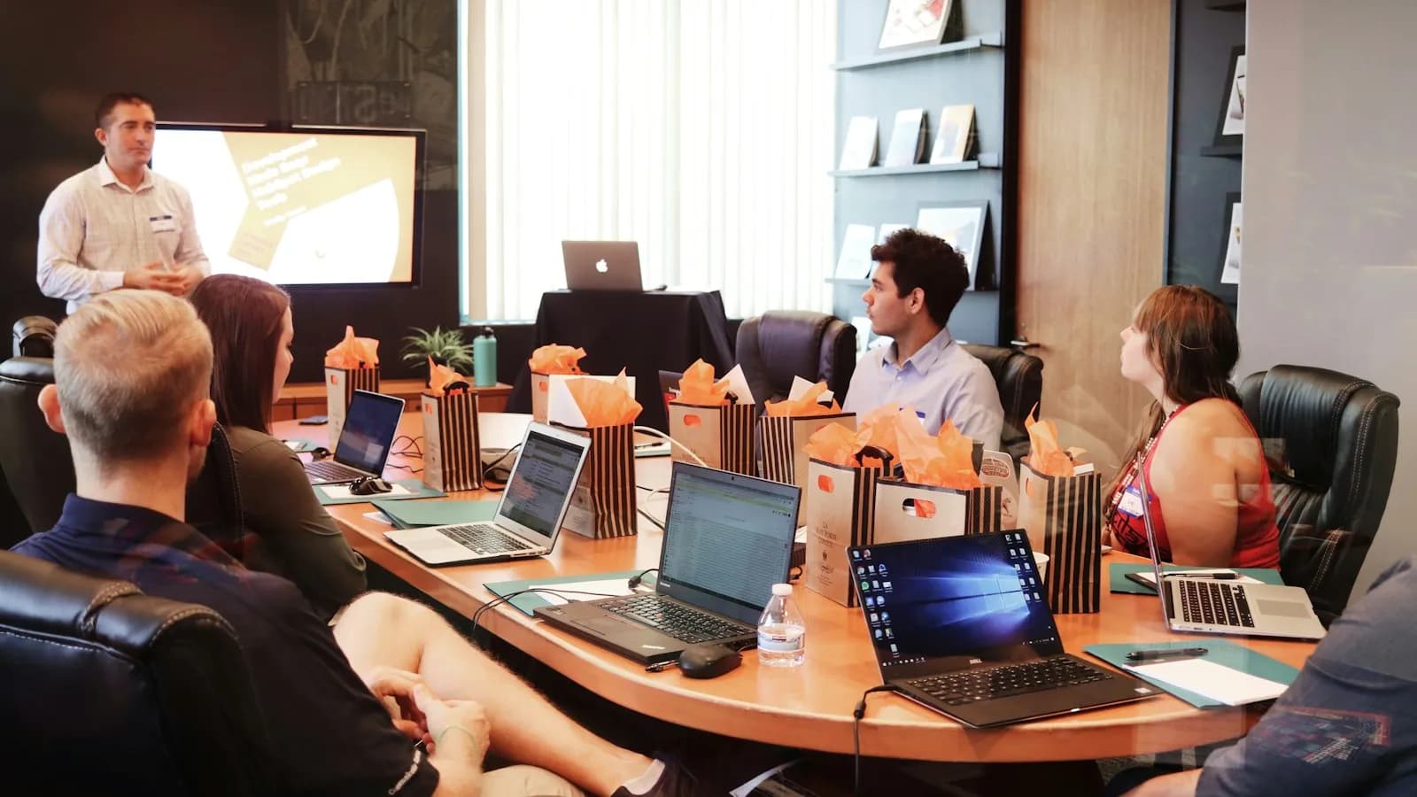 A man presenting to a team seated around a conference table with laptops