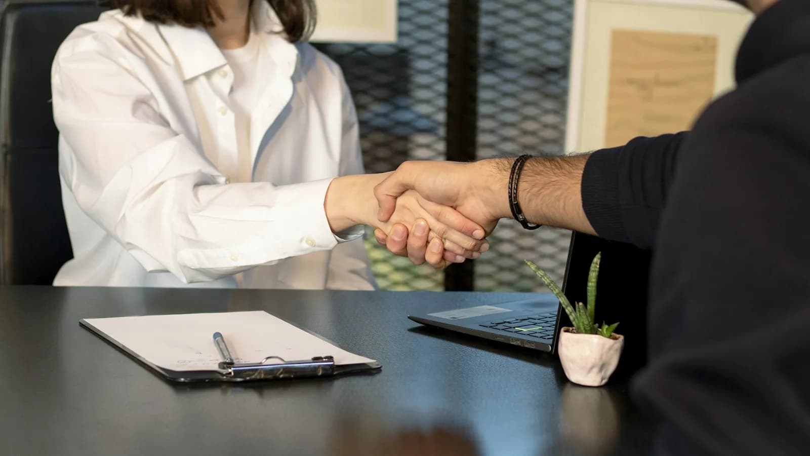 Two people shaking hands across a desk with a clipboard and laptop