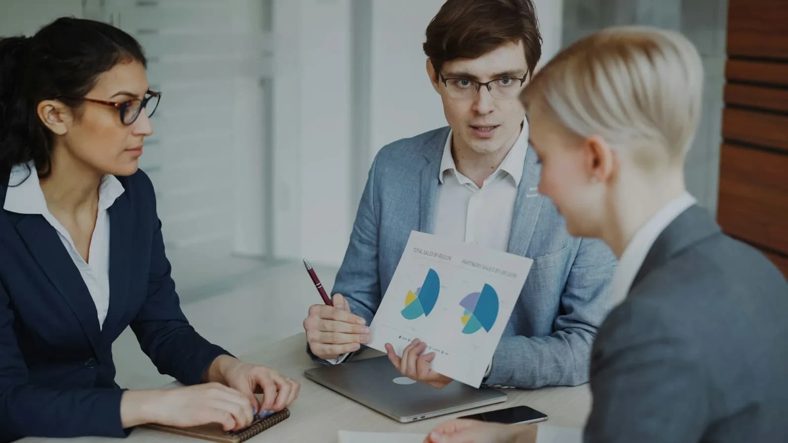 Three professionals at a table discussing printed charts, one holding a page with pie charts